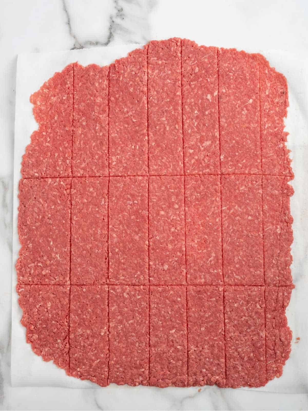 Raw ground beef is flattened into a large rectangle and scored into even square sections, resembling the convenient shape of Beef Jerky for Dogs, resting on a sheet of white parchment paper atop a marble surface.