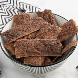 Bowl filled with homemade beef jerky strips for dogs on a white surface with a black and white patterned towel.