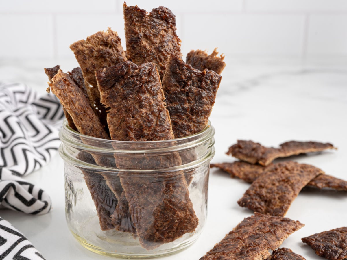 Beef jerky strips arranged upright in a glass jar, with more jerky pieces scattered on a white marble surface nearby. A black and white patterned cloth is partially visible on the left, highlighting this savory display of Beef Jerky for Dogs.