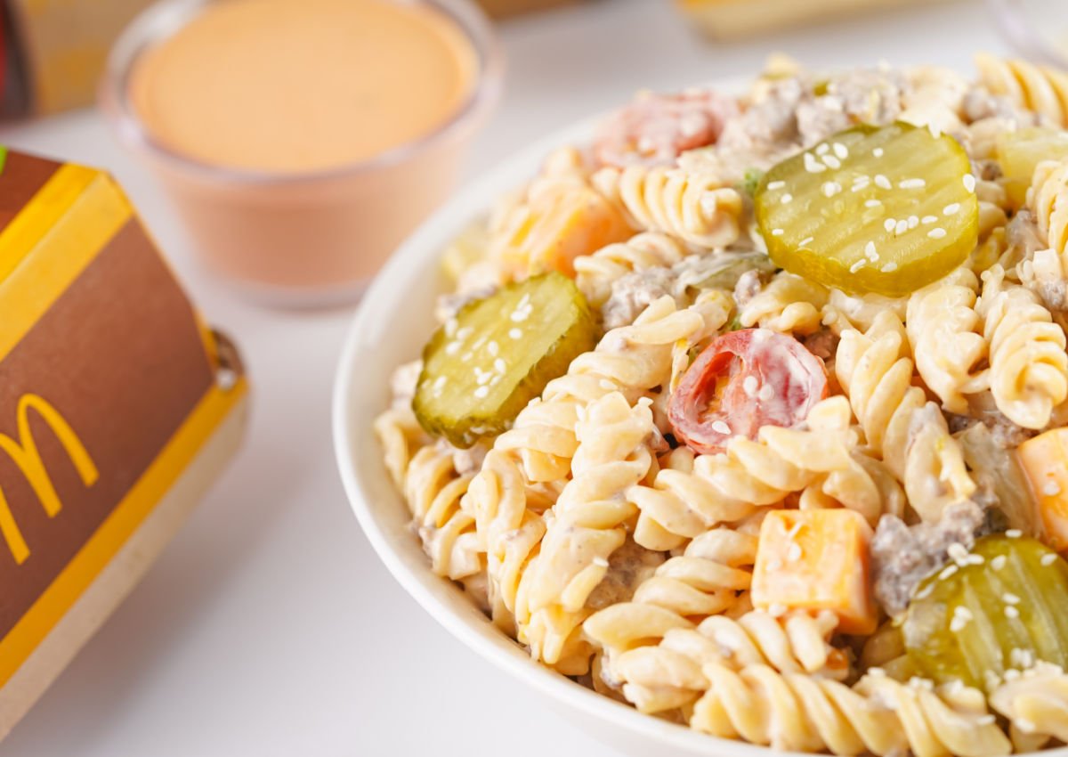 Close-up of Big Mac pasta salad in a white bowl with spiral pasta, pickles, ground beef, tomatoes, and cheddar cubes topped with sesame seeds.