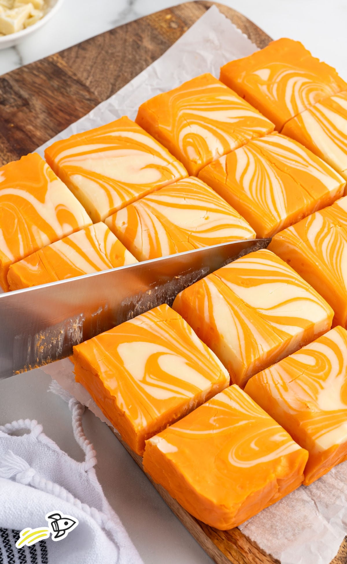 A close-up, high-angle shot of orange creamsicle fudge being sliced on a wooden cutting board lined with parchment paper. The fudge features a vibrant orange and creamy white swirled pattern. A large kitchen knife is positioned mid-cut through the neat, square blocks, revealing a dense and smooth texture. A white textured kitchen towel is visible in the bottom left corner.