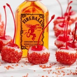 Close-up of bright red maraschino cherries dipped in white chocolate and red sugar with Fireball whiskey behind them.