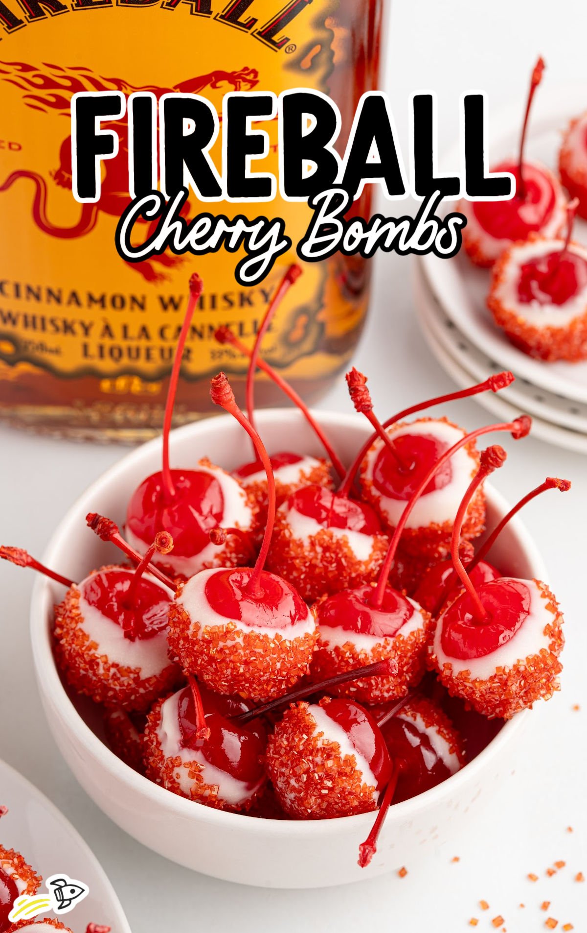 Hand holding a Fireball cherry bomb over stacked white plates beside a Fireball whiskey bottle.