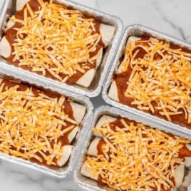 Four foil pans of freezer enchiladas covered in red sauce and shredded cheese on a marble countertop.