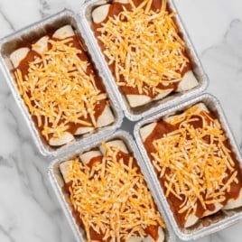 Four foil pans of prepared freezer enchiladas topped with red sauce and shredded cheese.