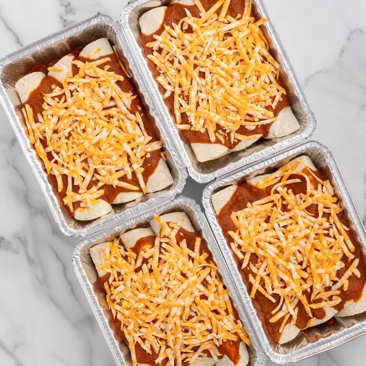 Four foil pans of prepared freezer enchiladas topped with red sauce and shredded cheese.