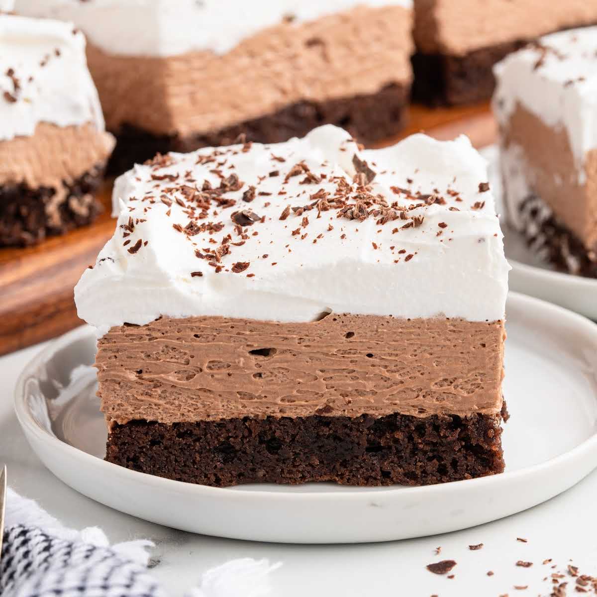 Single slice of french silk brownie on a white plate with visible layers of brownie base, chocolate mousse, and whipped cream, garnished with chocolate shavings.