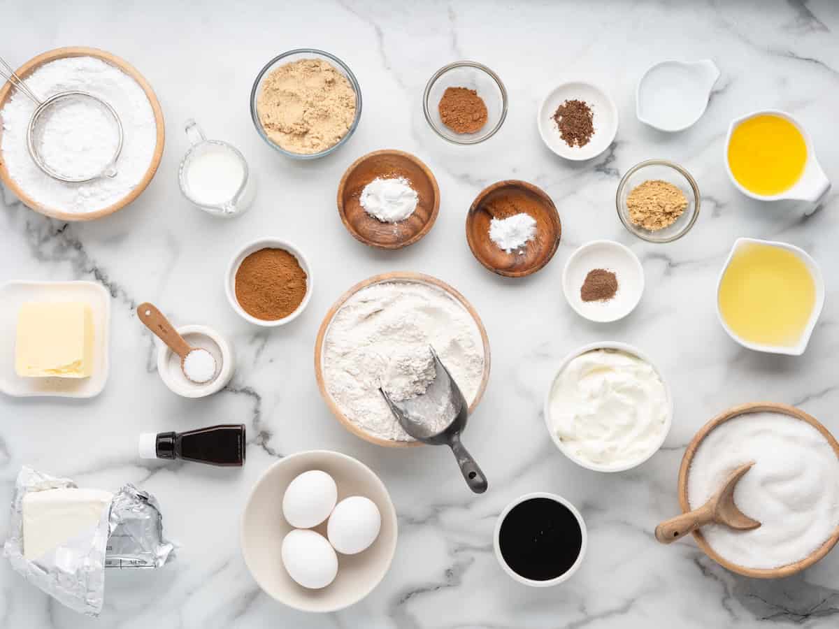 An overhead view of ingredients for a gingerbread cake arranged on a marble surface, including bowls of flour, sugar, brown sugar, eggs, butter, milk, oil, and various spices.