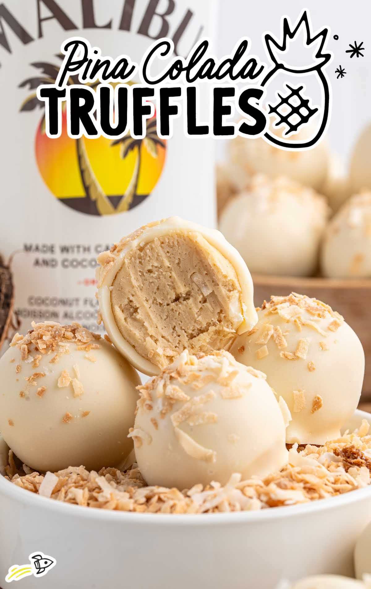 White chocolate pina colada truffles with toasted coconut topping stacked in a bowl in front of a Malibu rum bottle.