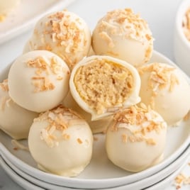 Pina colada truffles stacked on a white plate, coated in white chocolate and sprinkled with toasted coconut, with one truffle cut open to show filling.