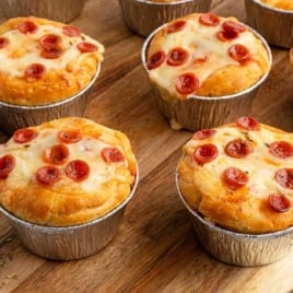 Freshly baked pizza pot pies in foil cups displayed on a wooden board with shredded mozzarella cheese and mini pepperoni on the side.
