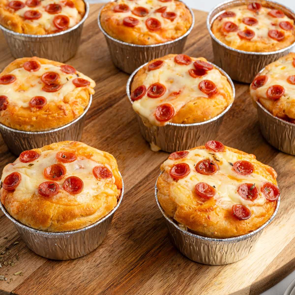 Close-up of baked pizza pot pies with melted cheese and pepperoni, arranged neatly on a wooden board in foil baking cups.