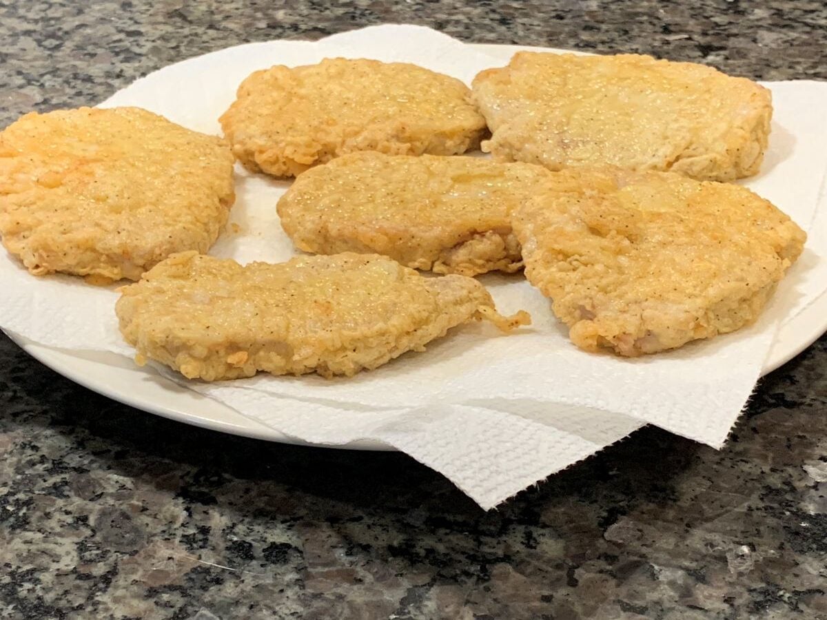 Six pieces of golden-brown fried chicken rest on paper towels atop a white plate on a granite countertop, inviting comparisons to the classic crunch of a Fried Pork Chops Recipe.