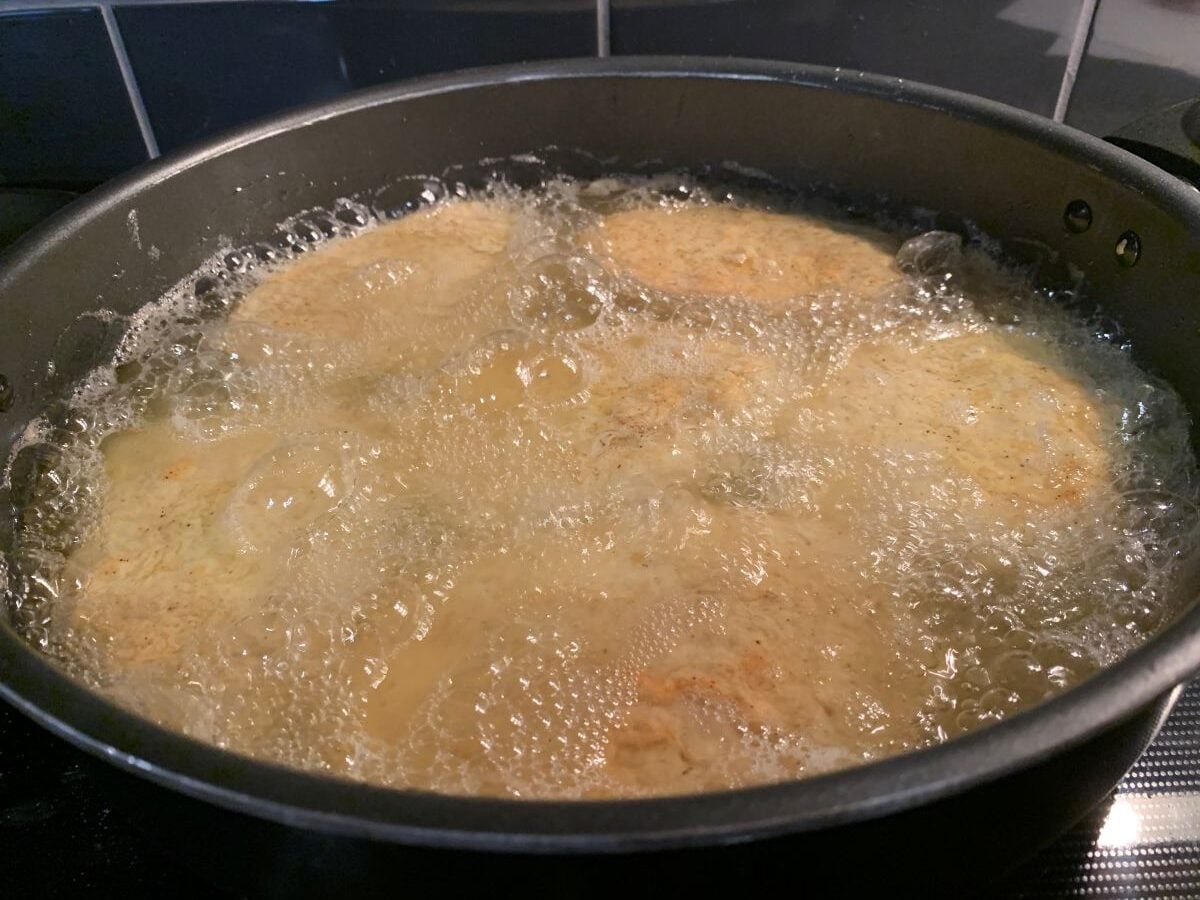 Four breaded patties, just like in a classic Fried Pork Chops Recipe, are sizzling in bubbling hot oil in a black frying pan on a stovetop, with a dark tiled backsplash visible in the background.