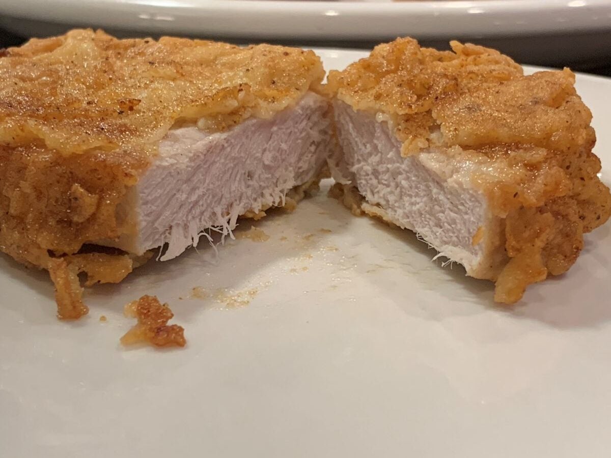 A close-up of a breaded and fried chicken breast cut in half, showing the crispy golden-brown coating and juicy white meat inside on a white plate, reminiscent of the classic crunch in a Fried Pork Chops Recipe.