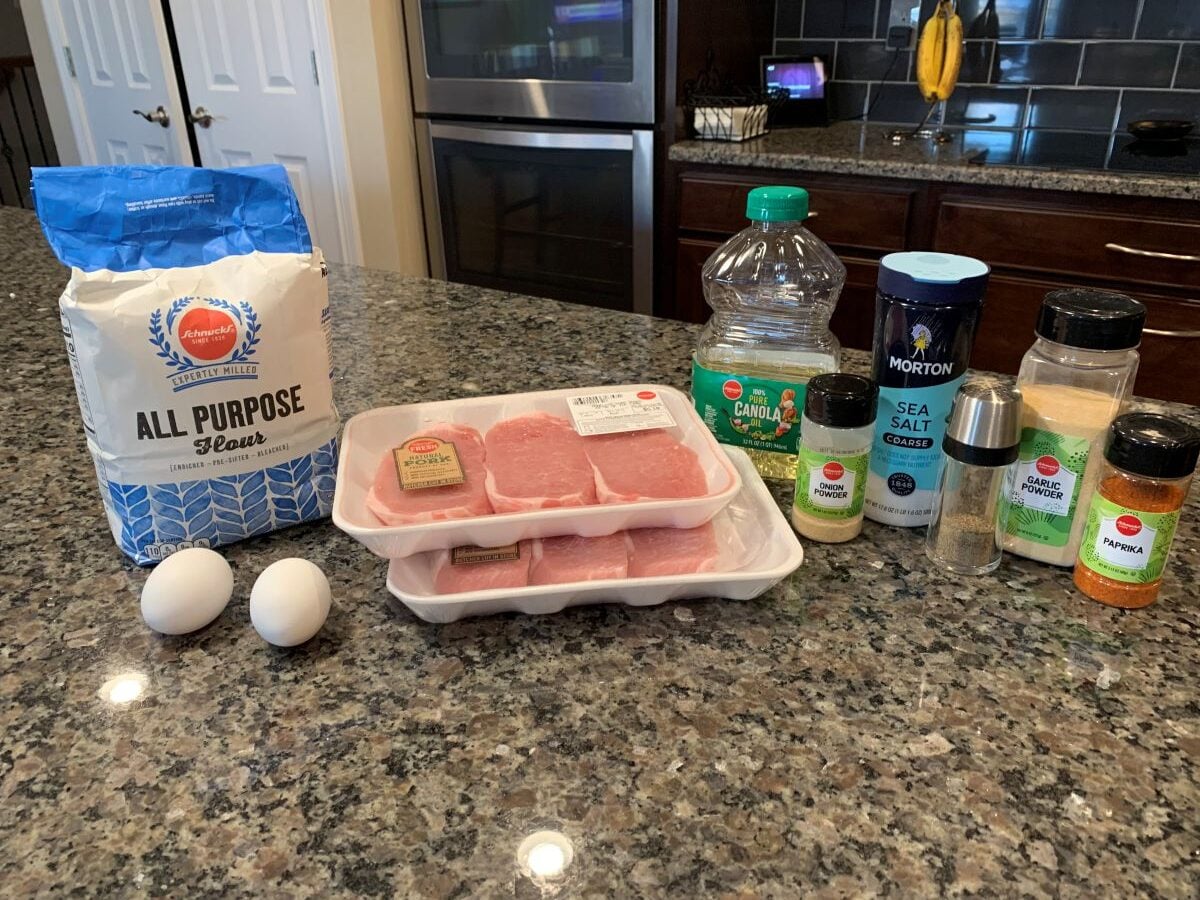 Ingredients for a Fried Pork Chops recipe on a kitchen counter: all-purpose flour, two eggs, two packs of pork chops, canola oil, sea salt, black pepper, garlic powder, onion powder, and paprika.