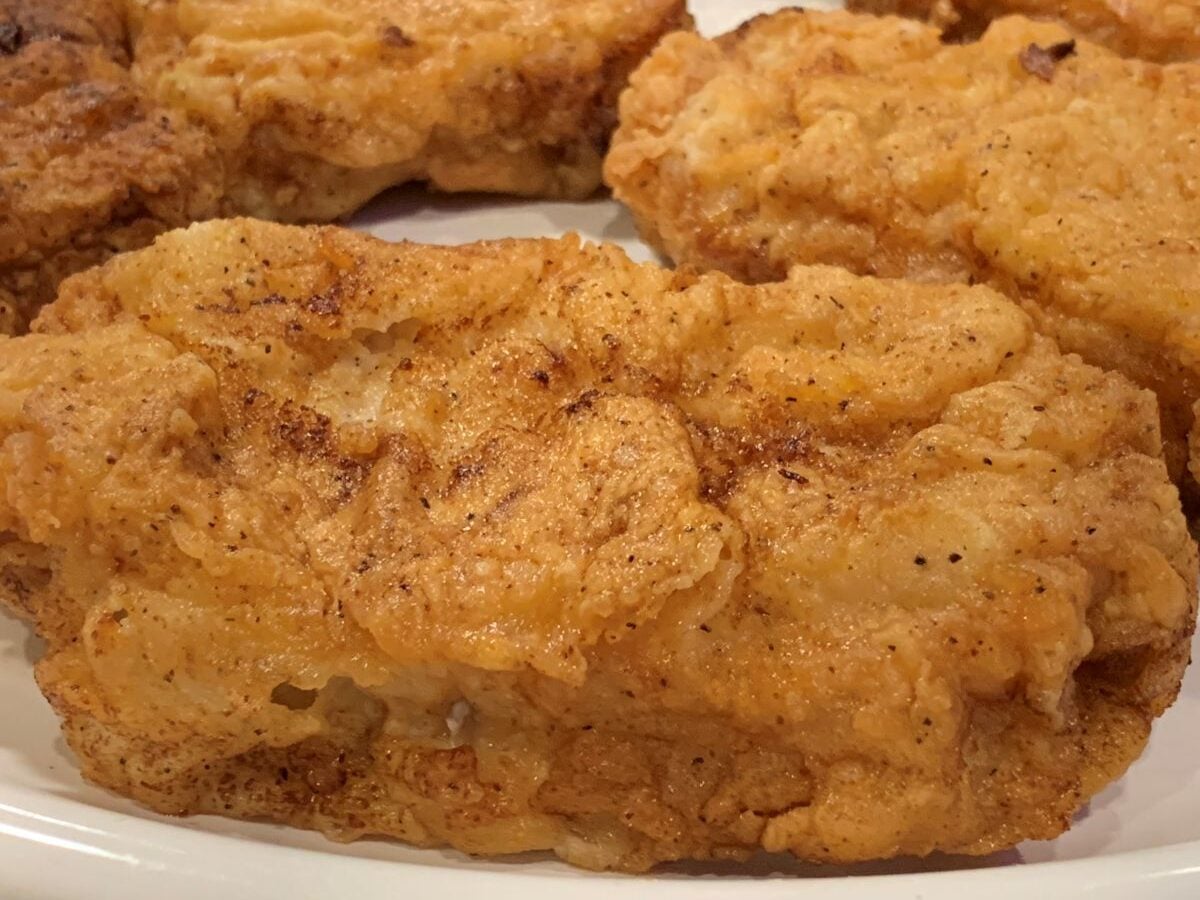 Close-up of golden-brown fried chicken with a crispy, textured coating, resting on a white plate—reminiscent of the crunch found in a classic Fried Pork Chops Recipe. Other pieces of fried chicken tempt in the background.