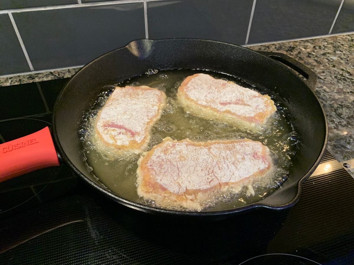 Three breaded pork chops sizzle in hot oil in a black cast iron skillet on the stovetop. The red silicone handle cover adds a pop of color, while this Fried Pork Chops Recipe fills the kitchen with irresistible aroma against a tiled backsplash.