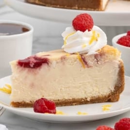 Slice of raspberry swirl lemon cheesecake on a white plate topped with whipped cream, raspberries, and lemon zest with coffee and fresh berries in the background.