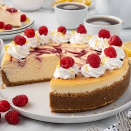 Whole raspberry swirl lemon cheesecake with a slice removed, decorated with whipped cream swirls, fresh raspberries, and lemon zest.