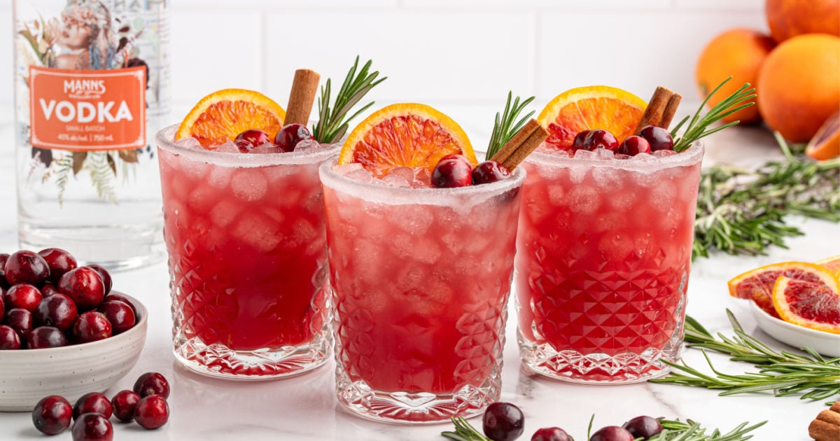 Three festive cranberry Christmas cocktails served in crystal glasses rimmed with sugar, each garnished with a slice of blood-orange, fresh cranberries, rosemary sprig, and a cinnamon stick, with a bottle of vodka and fresh ingredients in the background.