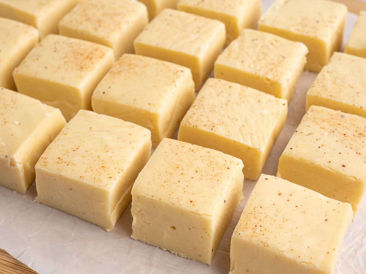Rows of creamy yellow Eggnog Fudge squares, each sprinkled lightly with spices, are arranged neatly on a sheet of parchment paper.