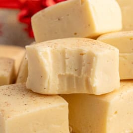 A close-up of creamy white Eggnog Fudge squares stacked together, with one piece showing a bite taken out of it. The fudge is lightly sprinkled with spices, capturing the festive flavors of this irresistible Eggnog Fudge Recipe.