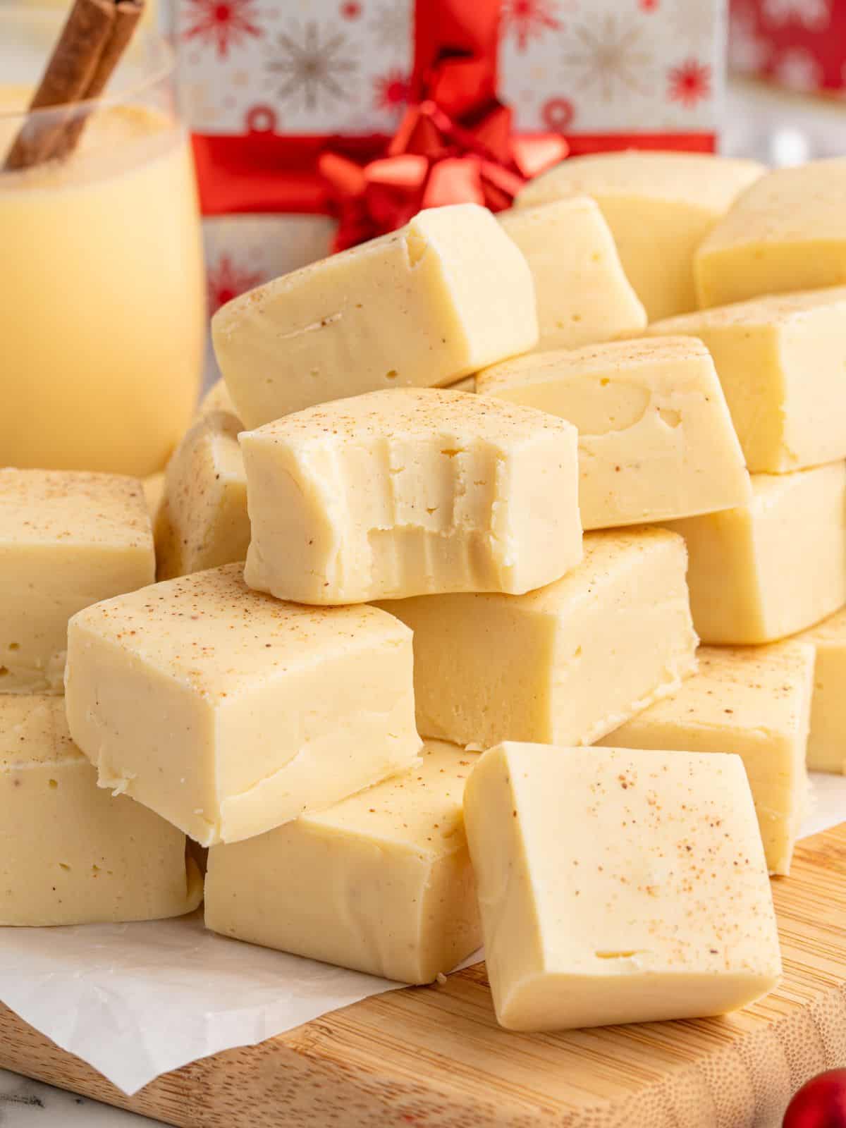 A stack of creamy white fudge squares, some dusted with spices, sits on a wooden board. One piece has bite marks, hinting at the irresistible appeal of this Eggnog Fudge Recipe. A glass of eggnog and a wrapped gift box are in the background.