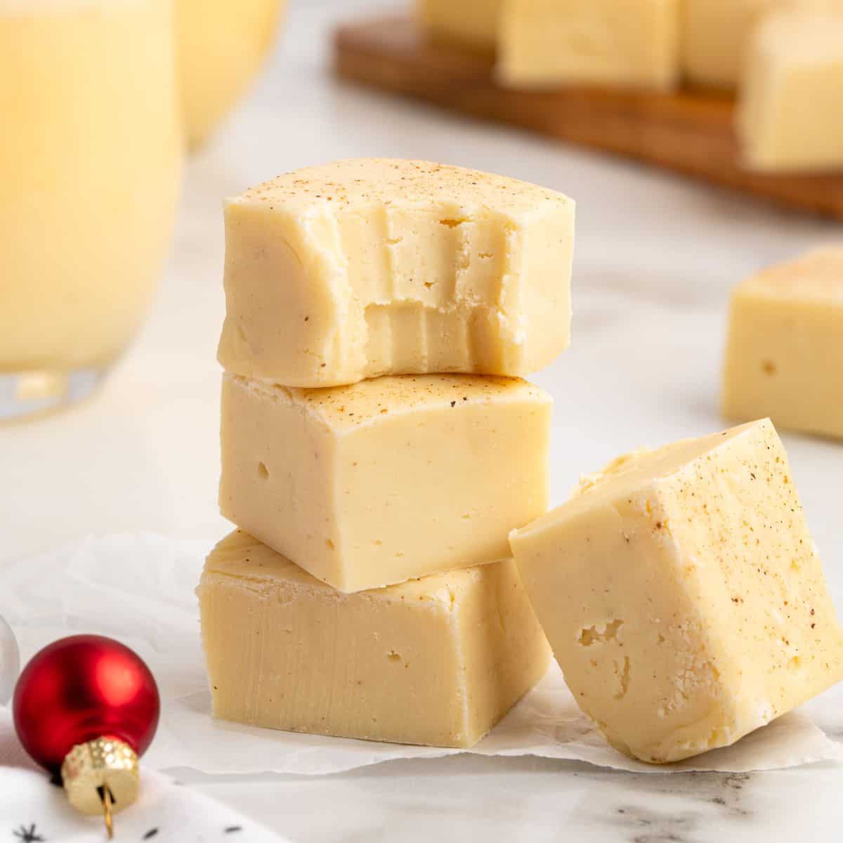 Four pieces of creamy eggnog fudge are stacked on parchment paper, with one piece showing bite marks—a delicious example of a classic Eggnog Fudge Recipe. A small red ornament and more fudge pieces are in the background, adding festive charm to this irresistible Eggnog Fudge treat.