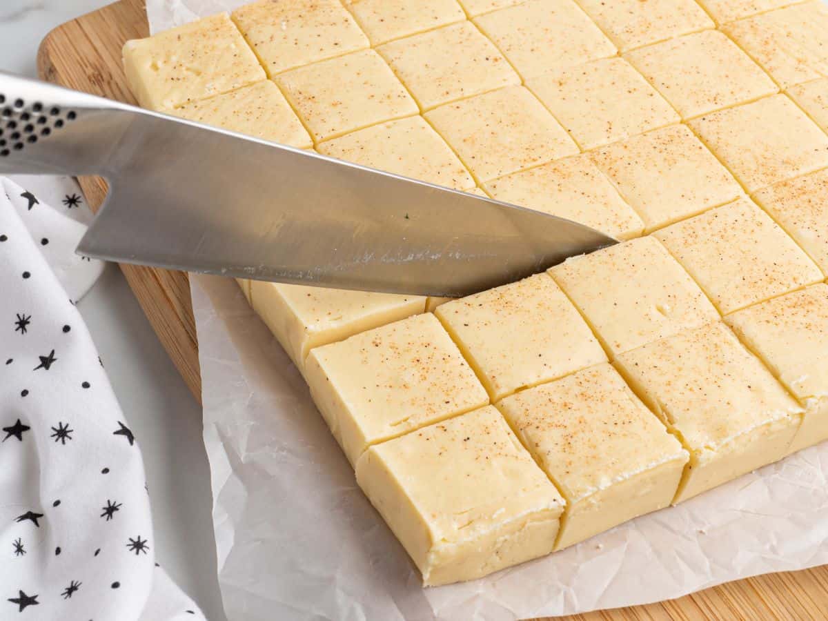 A large knife slices through a block of Eggnog Fudge cut into squares, resting on parchment paper on a wooden board. Some pieces are sprinkled with a light dusting of spice, adding a festive touch to this delicious Eggnog Fudge Recipe. A white cloth with black stars is nearby.