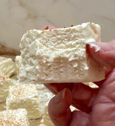 A hand holding a large, fluffy white marshmallow with a bite taken out. The background shows several more marshmallows on a light surface, reminiscent of the creamy sweetness found in an Eggnog Fudge recipe.