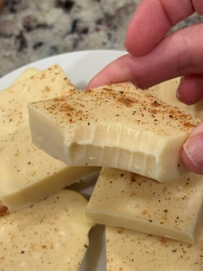 A hand holds a creamy white square dessert with a bite taken out, sprinkled with brown spice—an irresistible Eggnog Fudge treat. Several more pieces of this delectable Eggnog Fudge sit on a white plate in the background, ready to be enjoyed.