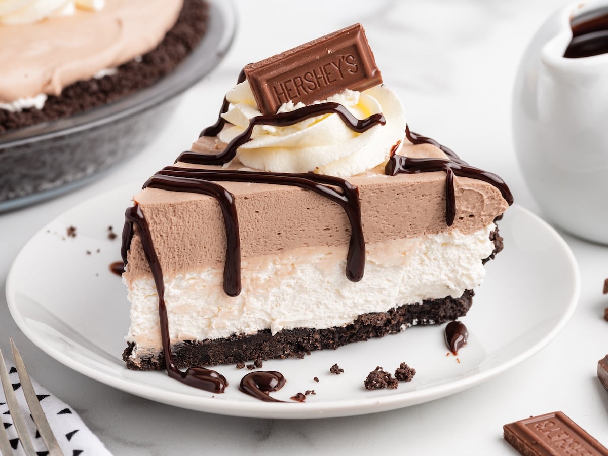 A slice of no-bake Hershey pie with chocolate drizzle, whipped cream, and a Hershey's chocolate bar on top, served on a white plate