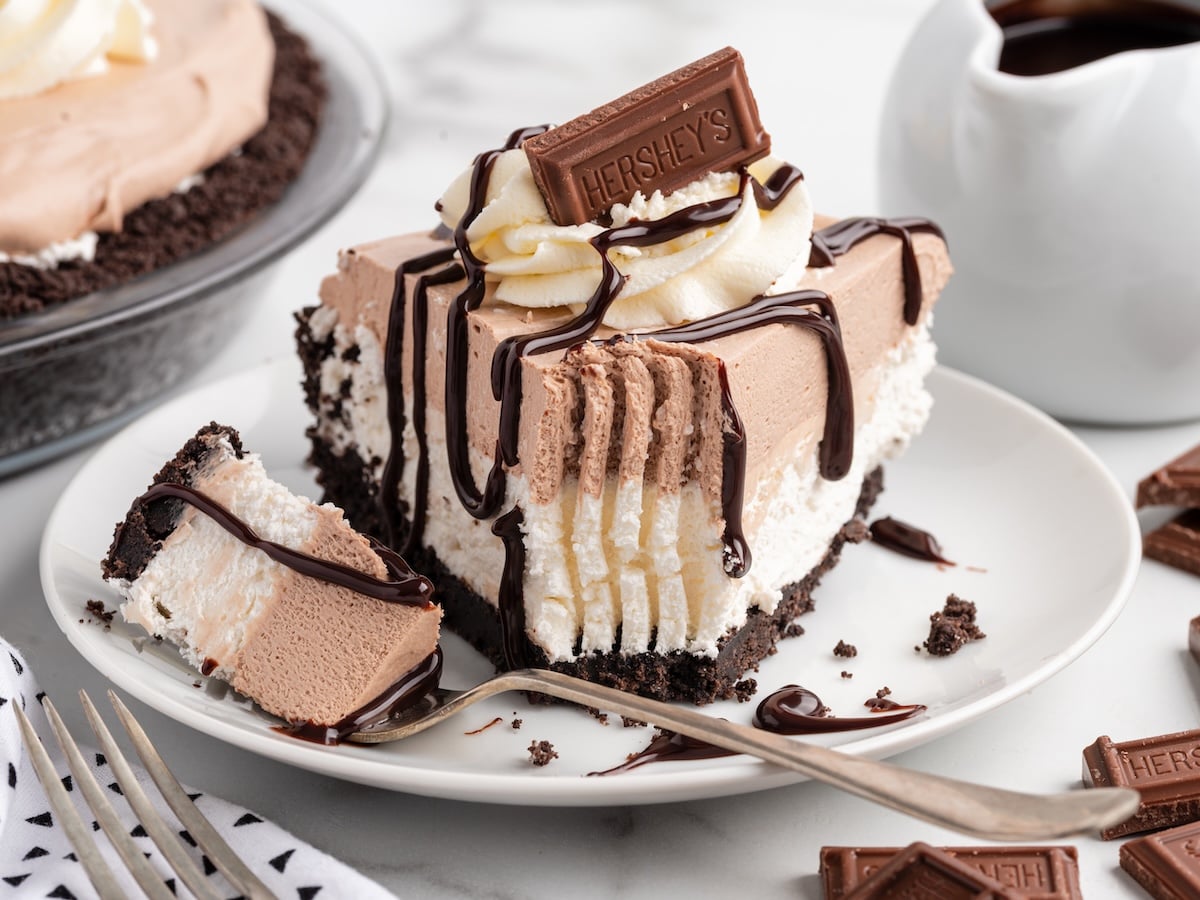 A slice of Hershey pie with layers of chocolate and cream filling in an Oreo crust, drizzled with chocolate sauce and topped with whipped cream and a Hershey's bar