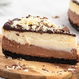 Triple chocolate cheesecake slice on a wooden board topped with chocolate ganache and shaved chocolate.