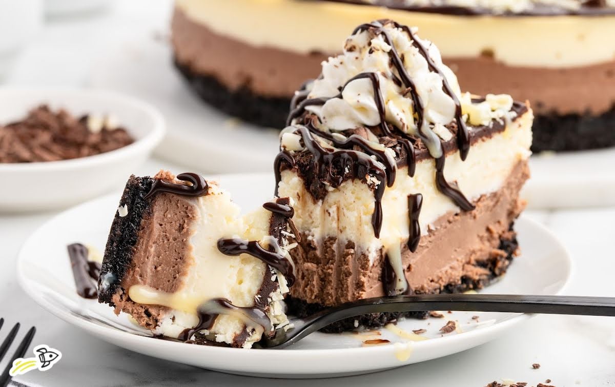 Triple chocolate cheesecake slice served with whipped cream, chocolate drizzle, and a forkful removed.