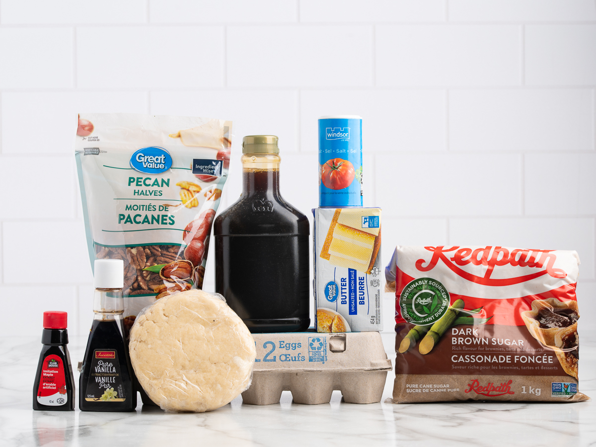 A variety of baking ingredients for a classic Pecan Pie Recipe, including pecan halves, eggs, butter, dark brown sugar, vanilla extract, pie dough, syrup, and whipped cream, arranged on a white marble countertop.