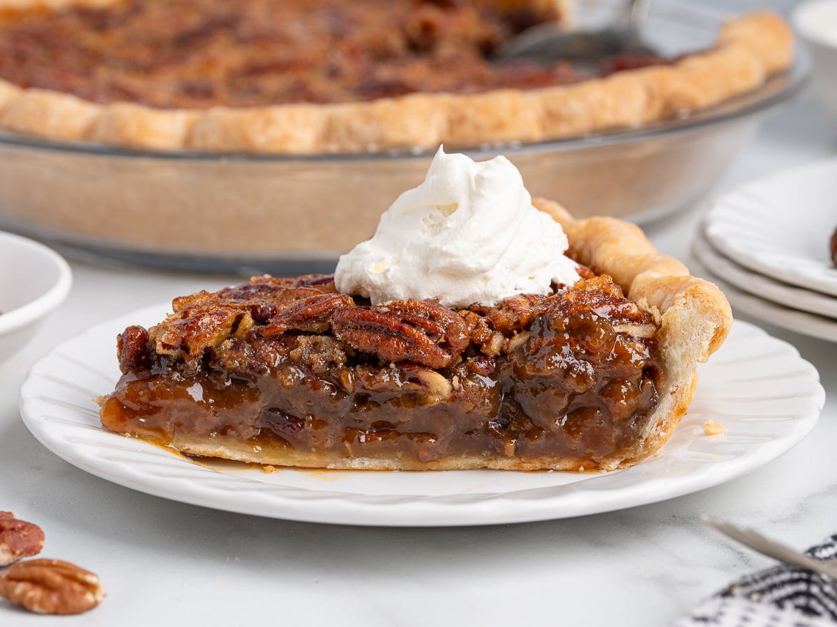 A slice of pecan pie, made from a classic pecan pie recipe, is topped with a dollop of whipped cream on a white plate, while the rest of the gooey, nut-packed dessert waits in the background.