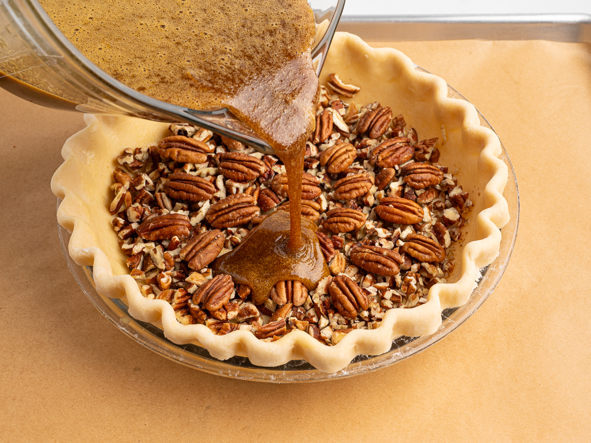 A glass bowl pours a brown, syrupy mixture over chopped and whole pecans in an unbaked crust, ready to be baked into a classic Pecan Pie Recipe, all set on a parchment-lined baking sheet.