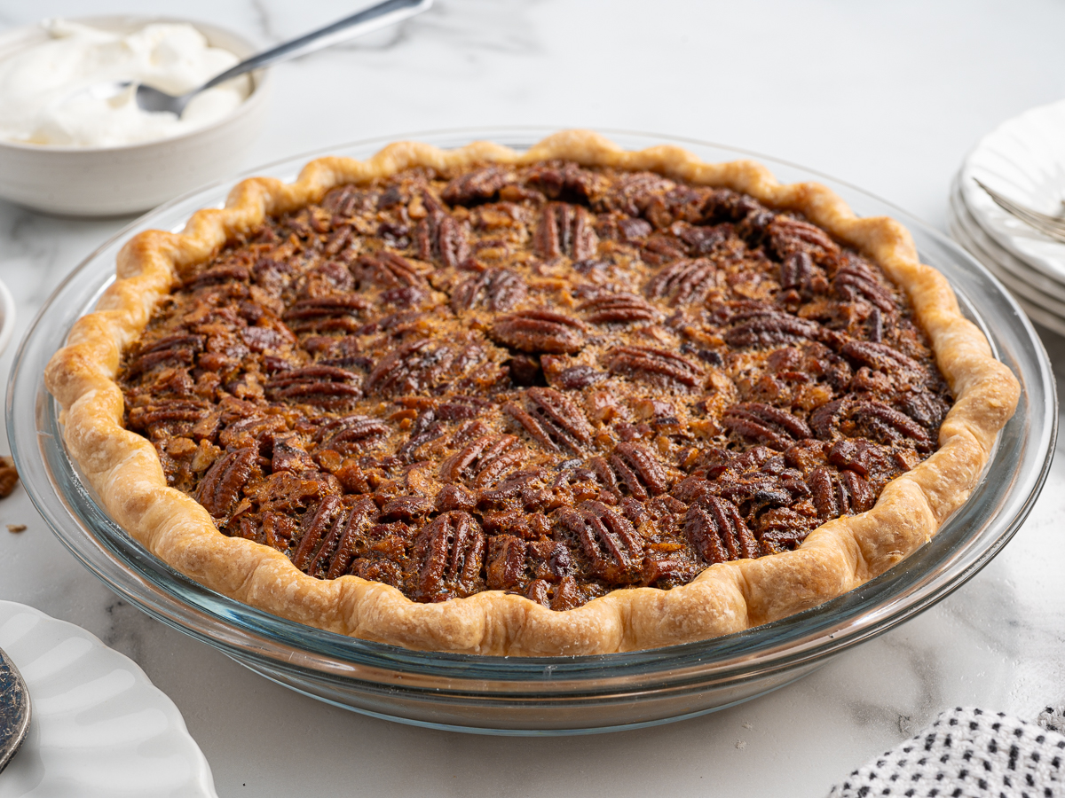 A pecan pie recipe with a golden, flaky crust sits in a glass pie dish. The surface is covered with whole pecans, and nearby are plates, a bowl of whipped cream, and a patterned cloth on a white surface.