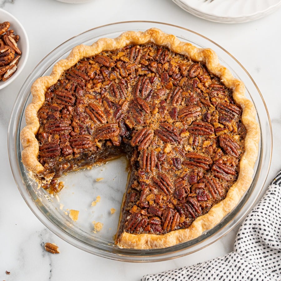 A pecan pie with a golden, flaky crust sits in a glass pie dish. One large slice has been removed, revealing the gooey filling and pecans insideโperfect inspiration for your next pecan pie recipe. A bowl of pecans and a dotted napkin are nearby.