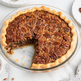A pecan pie made from a classic Pecan Pie Recipe, with a slice removed to reveal the gooey filling and pecan topping. The pie sits in a glass dish on marble, surrounded by plates, a bowl of pecans, and whipped cream.