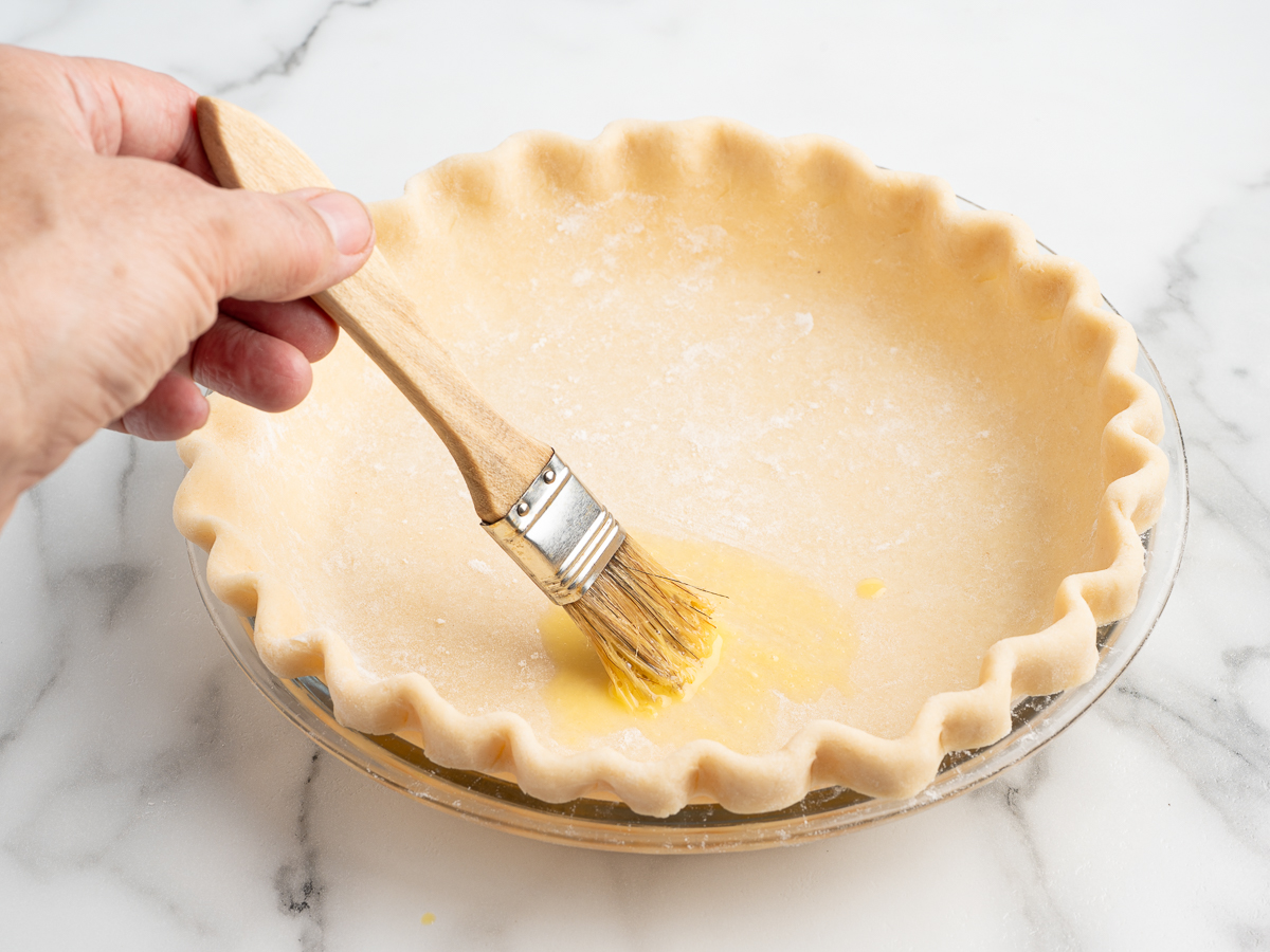 A hand uses a wooden pastry brush to apply an egg wash or butter to an unbaked, fluted pecan pie crust in a glass pie dish on a marble surface, preparing it for a delicious pecan pie recipe.