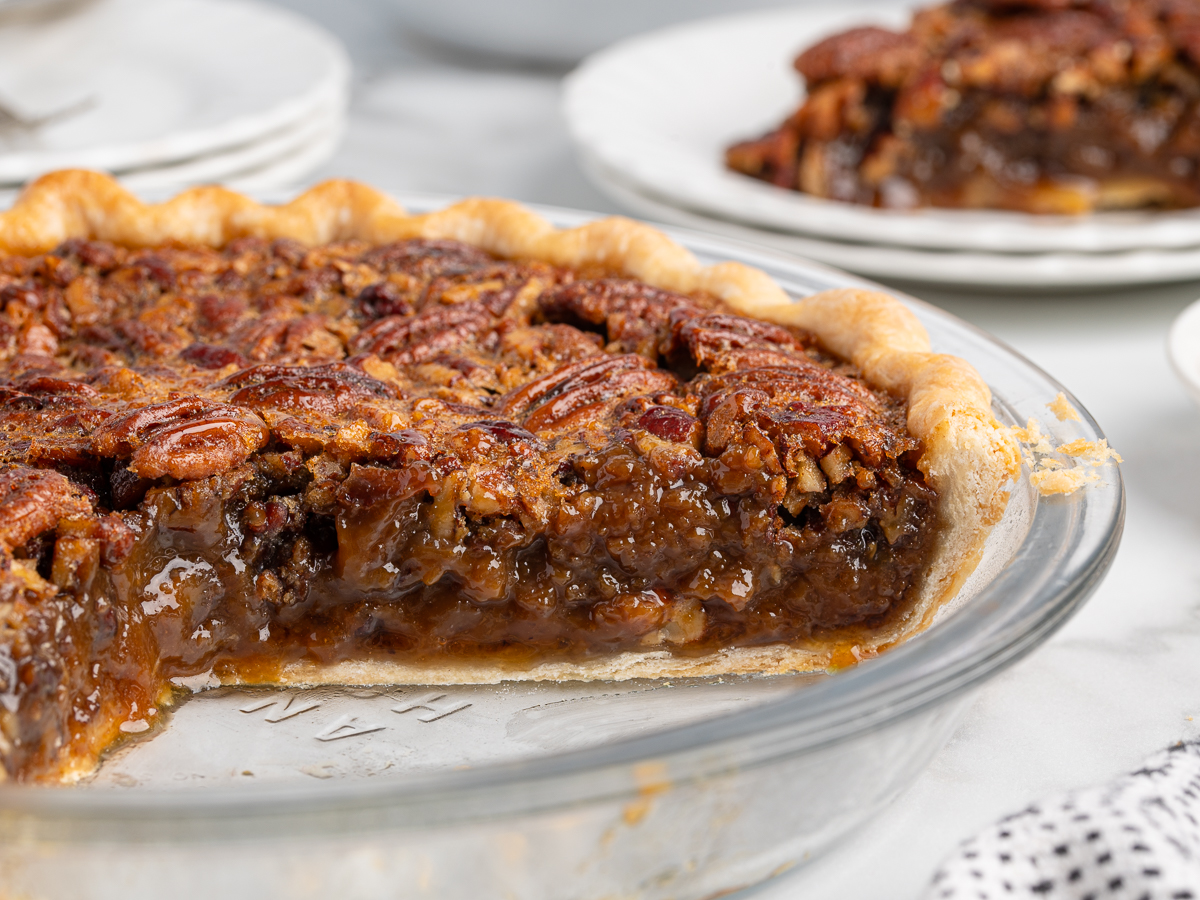 A close-up of a pecan pie in a glass pie dish with a slice missing, showing the gooey filling and pecan topping. In the background, a plated piece offers a glimpse of this irresistible pecan pie recipe.