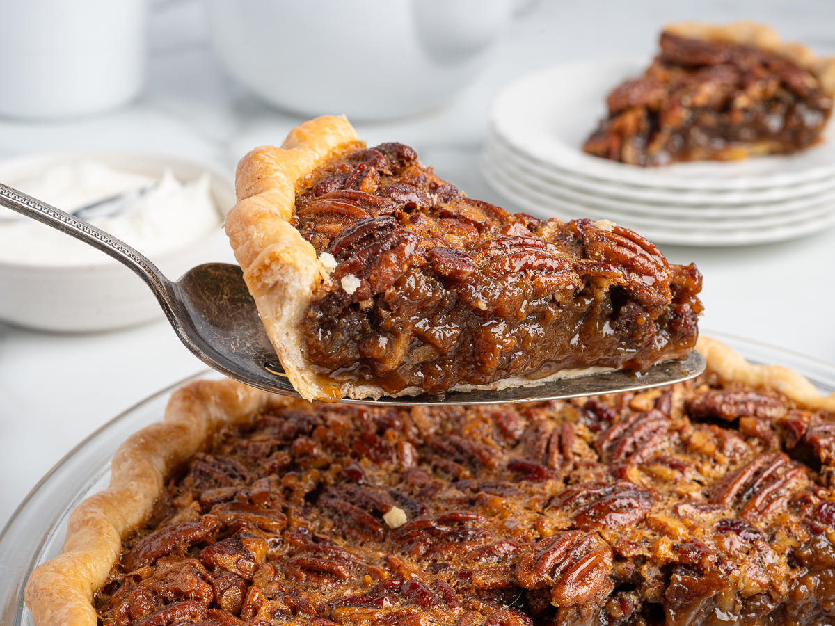 A close-up of a slice from a classic pecan pie recipe being lifted, highlighting the golden crust and glossy pecan filling. Plates with more pecan pie slices and a bowl of whipped cream appear in the background.