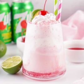 Glass of pink Shirley Temple float topped with whipped cream, a maraschino cherry, and a lime wedge with striped straws.