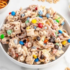 Close-up view of white chocolate snack mix filled with M&Ms, pretzels, Cheerios, and Chex cereal in a white bowl.