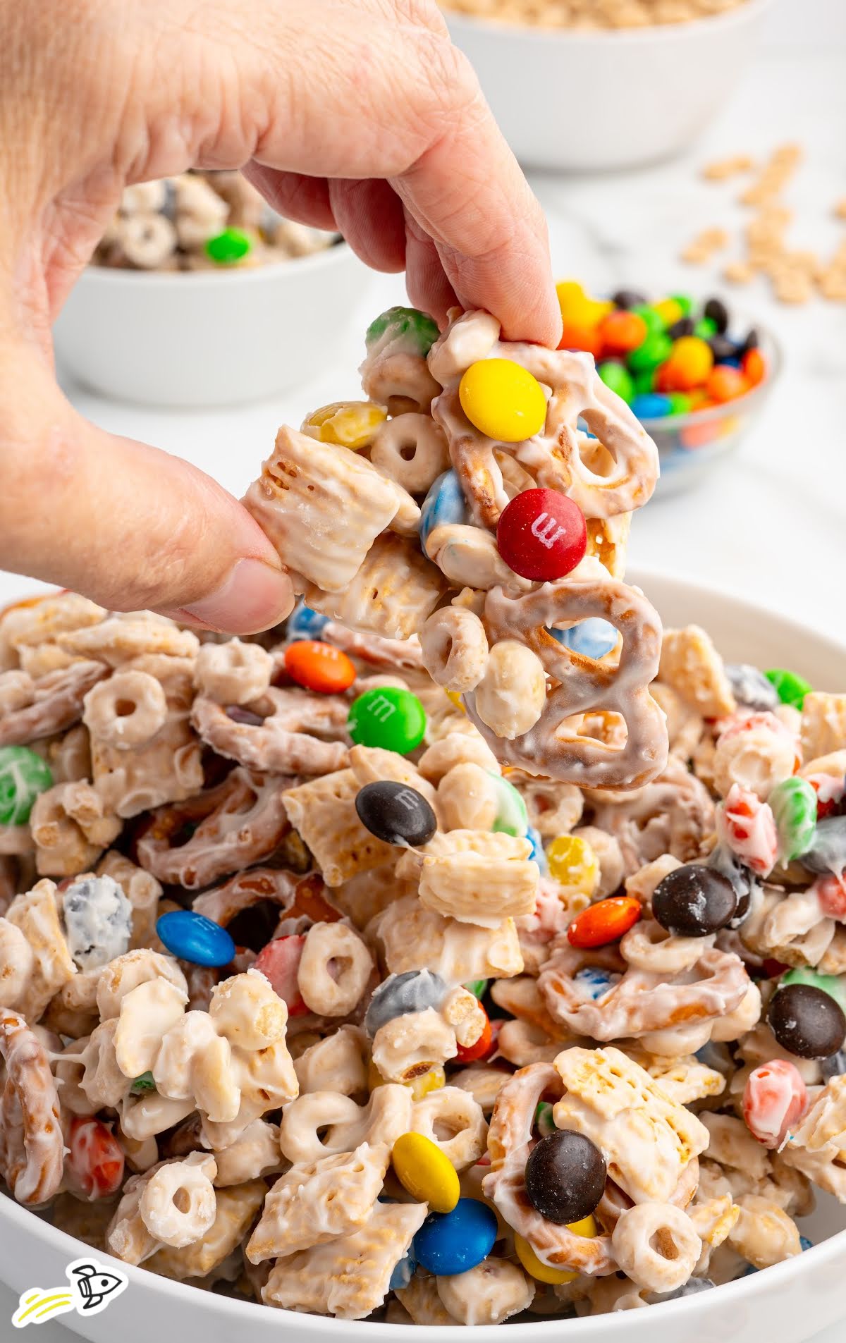 Hand lifting a cluster of white chocolate snack mix from a bowl, showing pieces of pretzels, M&Ms, and cereal covered in creamy coating.