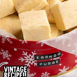 Squares of creamy Eggnog Fudge stacked in a festive red tin with snowflake patterns, topped with a handwritten label reading Eggnog Fudge and a sticker that says Vintage Recipe!! Perfect for sharing, this delightful treat is made using our cherished Eggnog Fudge Recipe.