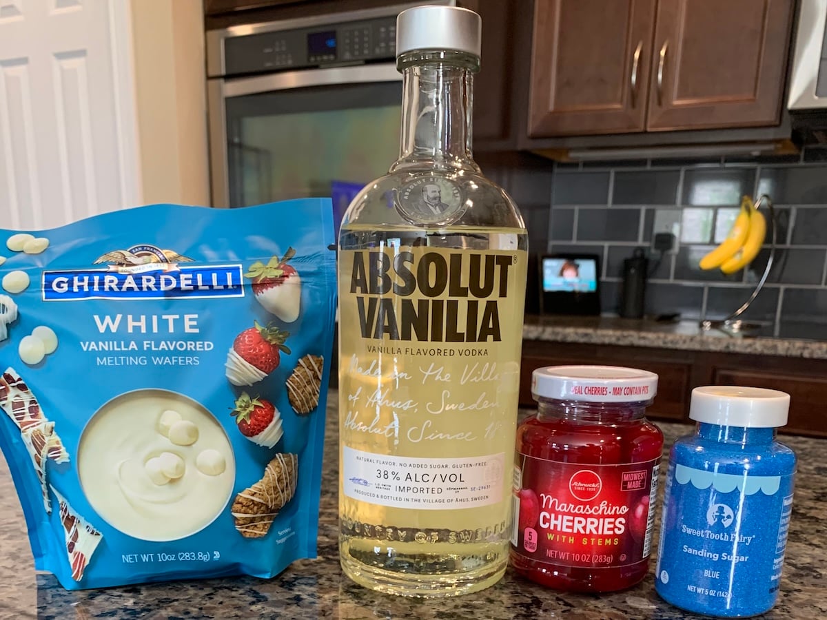 A bag of Ghirardelli white vanilla melting wafers, Absolut Vanilia vodka, maraschino cherries, and blue sprinkles are displayed on the counter&mdash;perfect ingredients for making festive 4th of July Cherry Bombs.